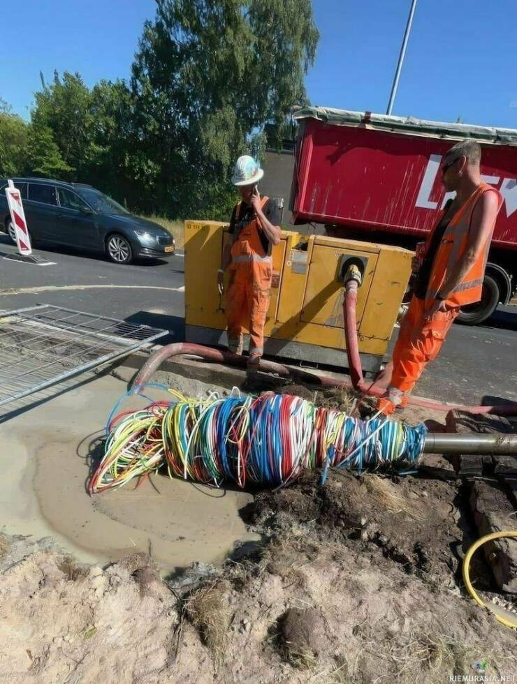 Construction workers digging a hole next to a road using a vertical boring machine. The drill bit, around 10-15cm in diameter, has hit a big wire bundle and wound up lots of wires, like a fork in spaghetti